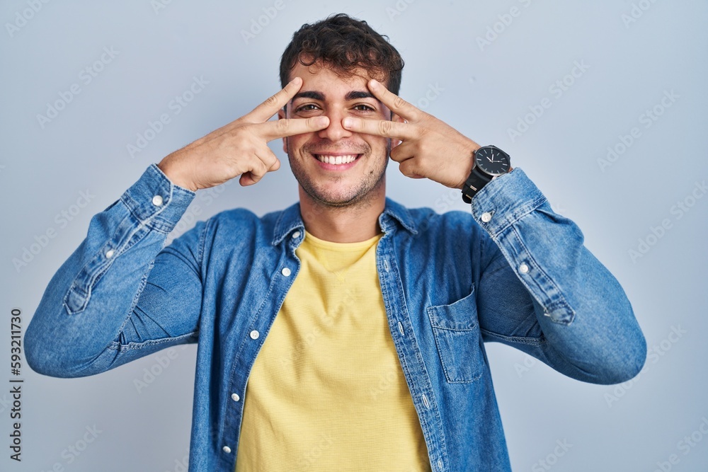 Young hispanic man standing over blue background doing peace symbol with fingers over face, smiling cheerful showing victory Young hispanic man standing over blue background doing peace symbol with fingers over face, smiling cheerful showing victory