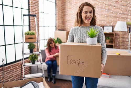 Mother and daughter moving to a new home holding cardboard box winking looking at the camera with sexy expression, cheerful and happy face. © Krakenimages.com