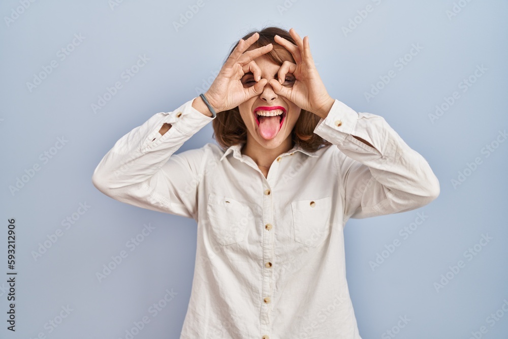 Young beautiful woman standing casual over blue background doing ok gesture like binoculars sticking tongue out, eyes looking through fingers. crazy expression. Young beautiful woman standing casual over blue background doing ok gesture like binoculars sticking tongue out, eyes looking through fingers. crazy expression.