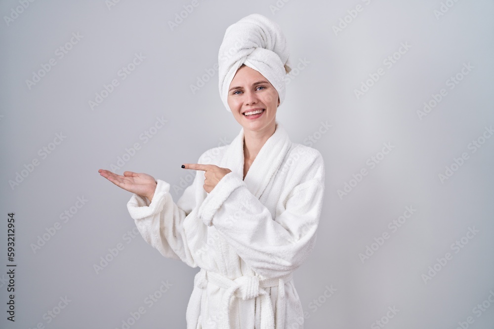 Blonde caucasian woman wearing bathrobe amazed and smiling to the camera while presenting with hand and pointing with finger.