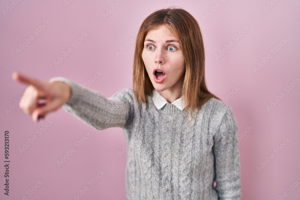 Beautiful woman standing over pink background pointing with finger surprised ahead, open mouth amazed expression, something on the front Beautiful woman standing over pink background pointing with finger surprised ahead, open mouth amazed expression, something on the front
