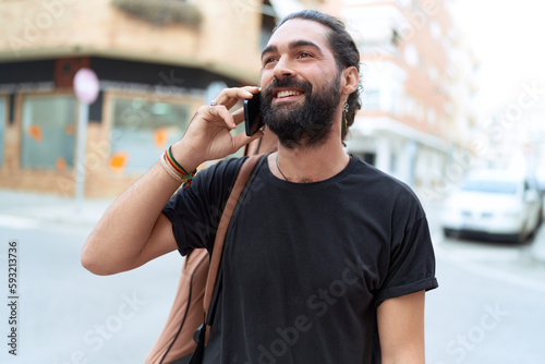 Young hispanic man musician talking on smartphone holding guitar case at street