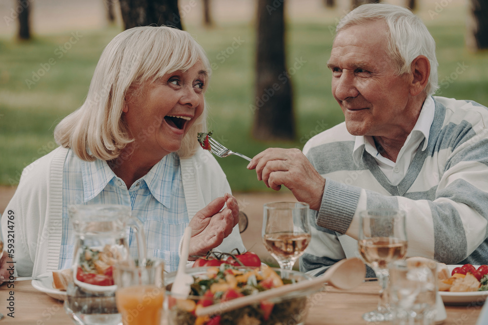 Cheerful senior couple feeding each other while having dinner outdoors together Cheerful senior couple feeding each other while having dinner outdoors together