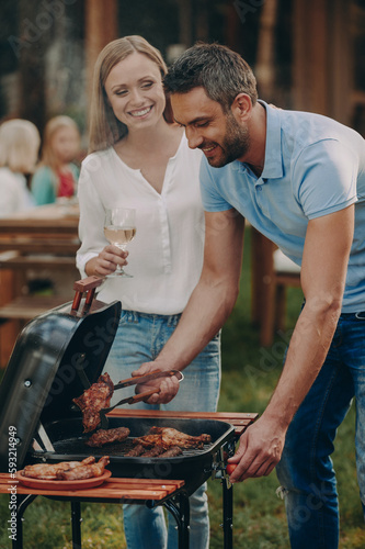Happy young couple barbecuing meat on grill while their family relaxing in the background
