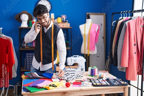 Young hispanic man tailor talking on smartphone writing on notebook at sewing studio