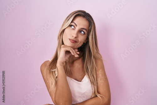 Young blonde woman standing over pink background with hand on chin thinking about question, pensive expression. smiling with thoughtful face. doubt concept.
