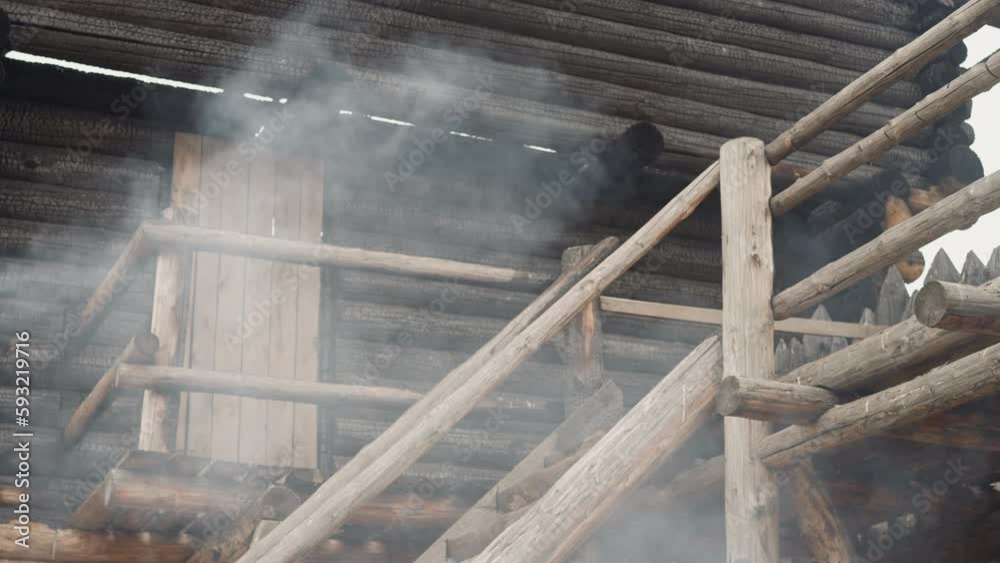 White smoke from bonfire billows near high log cabin. Traditional medieval wooden dwelling with stairs. Cooking food on fire in yard on warm day White smoke from bonfire billows near high log cabin. Traditional medieval wooden dwelling with stairs. Cooking food on fire in yard on warm day