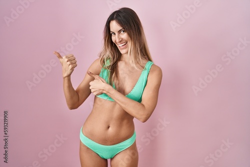 Young hispanic woman wearing bikini over pink background pointing to the back behind with hand and thumbs up, smiling confident