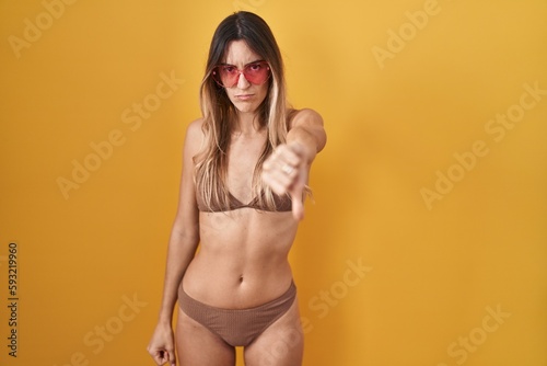 Young hispanic woman wearing bikini over yellow background looking unhappy and angry showing rejection and negative with thumbs down gesture. bad expression.