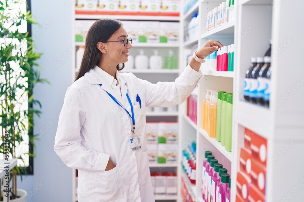Young beautiful hispanic woman pharmacist smiling confident holding product on shelving at pharmacy