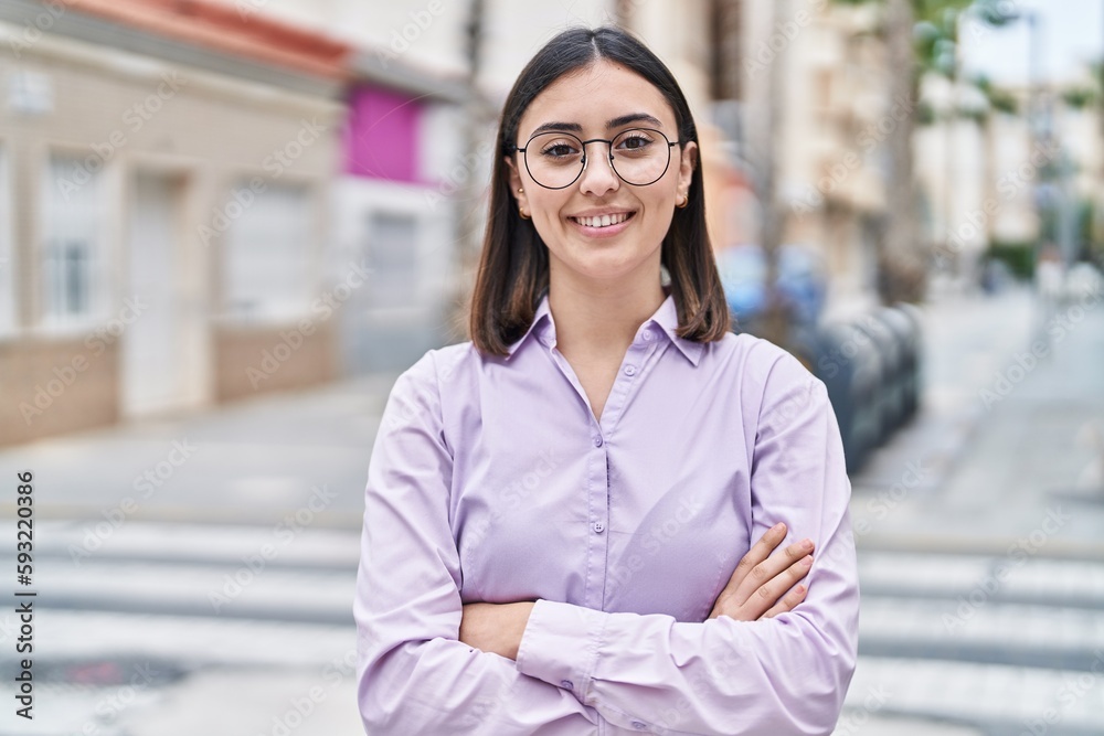 Young hispanic woman standing with relaxed expression and arms crossed gesture at street
