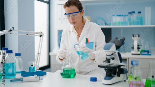 Middle age hispanic woman wearing scientist uniform pouring liquid on test tube at laboratory