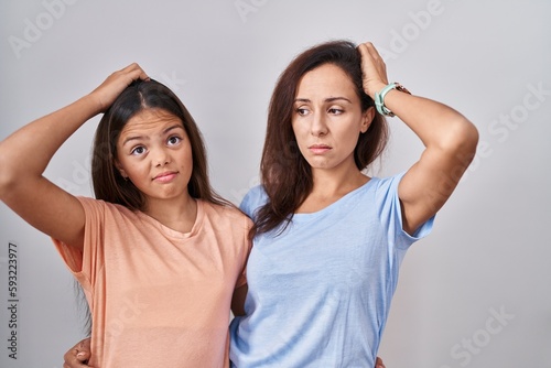 Young mother and daughter standing over white background confuse and wondering about question. uncertain with doubt  thinking with hand on head. pensive concept.