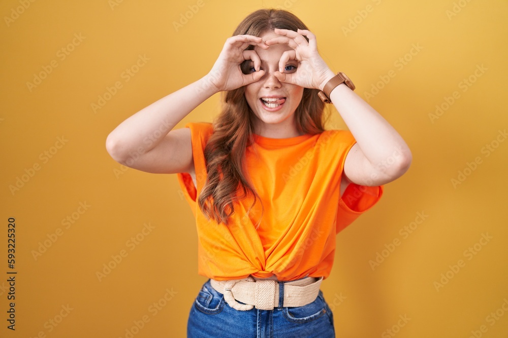 Caucasian woman standing over yellow background doing ok gesture like binoculars sticking tongue out, eyes looking through fingers. crazy expression.