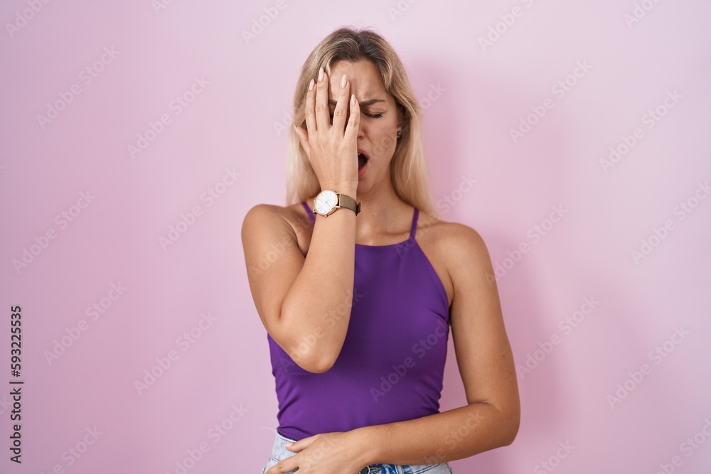 Young blonde woman standing over pink background yawning tired covering half face, eye and mouth with hand. face hurts in pain. Young blonde woman standing over pink background yawning tired covering half face, eye and mouth with hand. face hurts in pain.