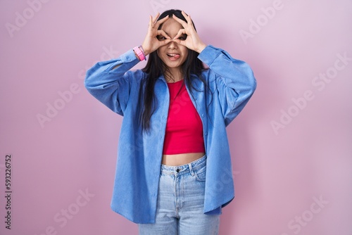 Young asian woman standing over pink background doing ok gesture like binoculars sticking tongue out, eyes looking through fingers. crazy expression. © Krakenimages.com