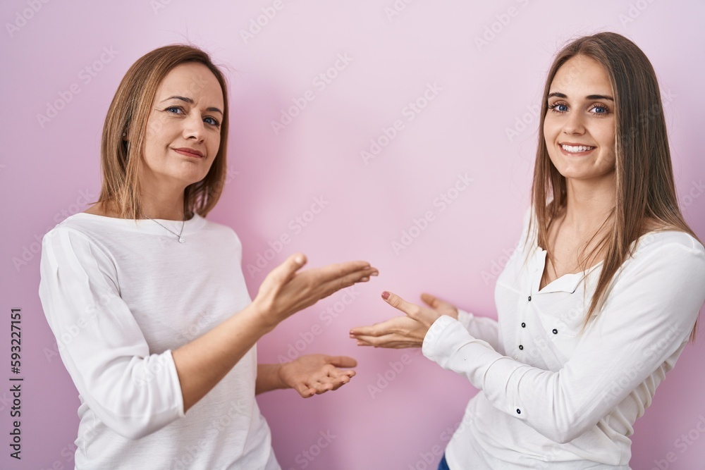 Middle age mother and young daughter standing over pink background inviting to enter smiling natural with open hand Middle age mother and young daughter standing over pink background inviting to enter smiling natural with open hand