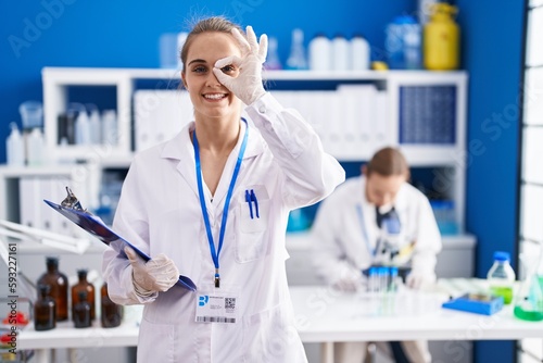 Blonde woman working at scientist laboratory smiling happy doing ok sign with hand on eye looking through fingers