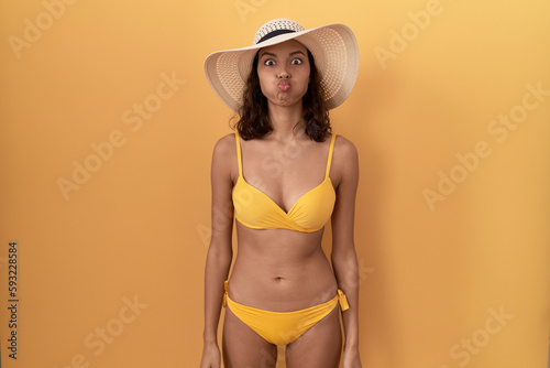 Fényképezés Young hispanic woman wearing bikini and summer hat puffing cheeks with funny face