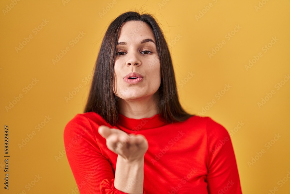 Young hispanic woman standing over yellow background looking at the camera blowing a kiss with hand on air being lovely and sexy. love expression. Young hispanic woman standing over yellow background looking at the camera blowing a kiss with hand on air being lovely and sexy. love expression.