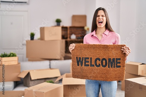 Young hispanic woman holding welcome doormat at new home angry and mad screaming frustrated and furious, shouting with anger looking up. © Krakenimages.com