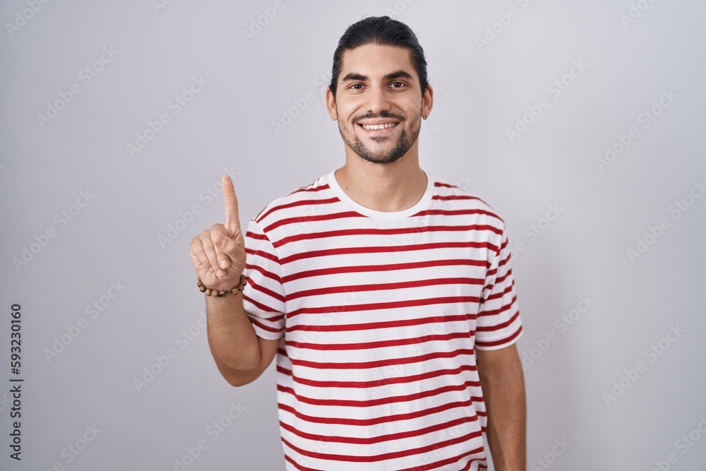 Hispanic man with long hair standing over isolated background showing and pointing up with finger number one while smiling confident and happy. Hispanic man with long hair standing over isolated background showing and pointing up with finger number one while smiling confident and happy.