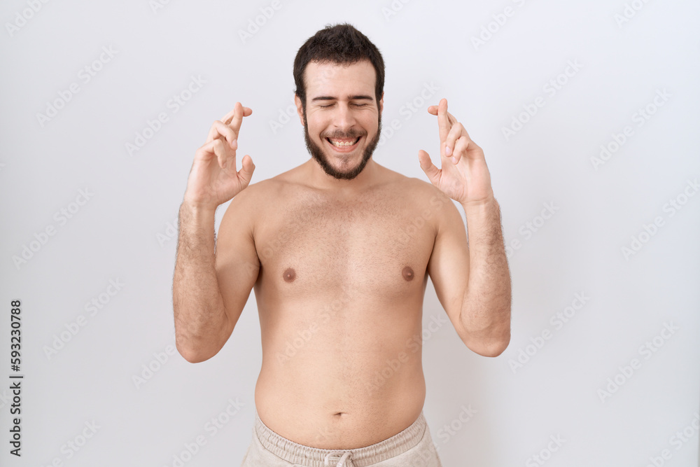 Young hispanic man standing shirtless over white background gesturing finger crossed smiling with hope and eyes closed. luck and superstitious concept.