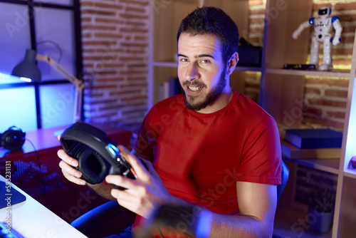 Young hispanic man streamers sitting on table holding headphones at gaming room