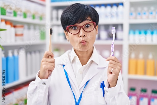 Young asian woman with short hair doing toothbrush comparative at pharmacy making fish face with mouth and squinting eyes, crazy and comical.