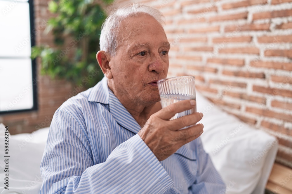 Middle age grey-haired man drinking glass of water sitting on bed at bedroom Middle age grey-haired man drinking glass of water sitting on bed at bedroom