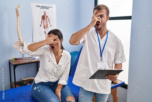 Young hispanic woman at physiotherapist appointment peeking in shock covering face and eyes with hand, looking through fingers with embarrassed expression. photo