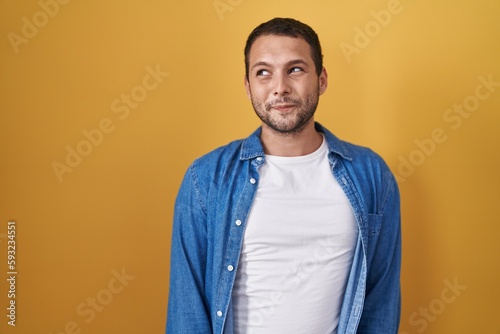 Hispanic man standing over yellow background smiling looking to the side and staring away thinking.