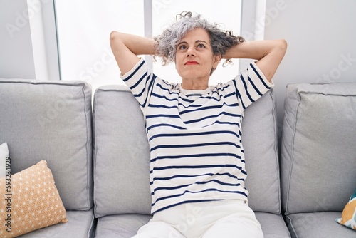 Middle age grey-haired woman relaxed with hands on head sitting on sofa at home