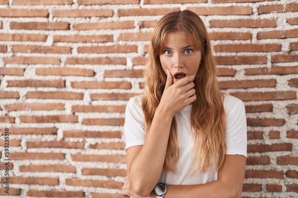 Young caucasian woman standing over bricks wall looking fascinated with disbelief, surprise and amazed expression with hands on chin