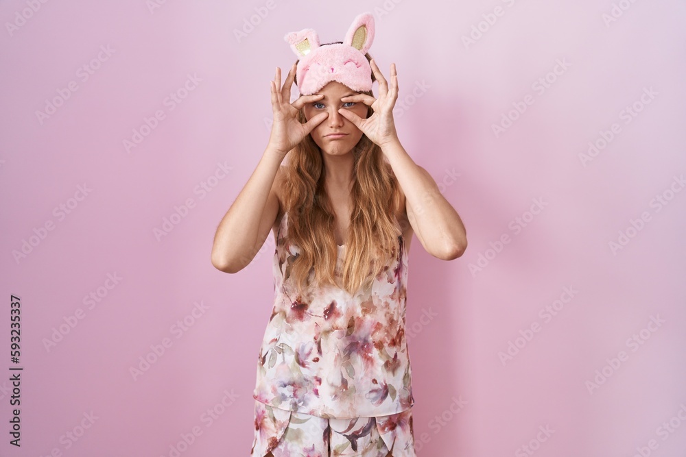Young caucasian woman wearing sleep mask and pajama trying to open eyes with fingers, sleepy and tired for morning fatigue