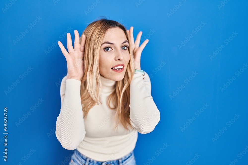 Young caucasian woman standing over blue background trying to hear both hands on ear gesture, curious for gossip. hearing problem, deaf