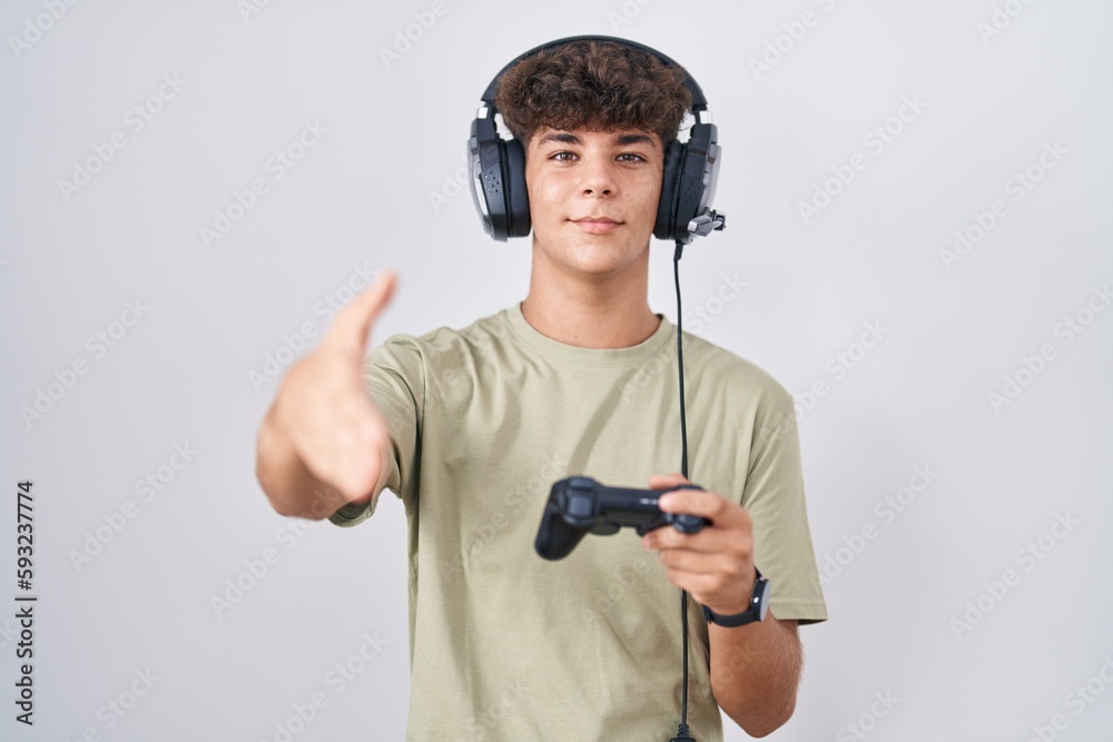 Hispanic teenager playing video game holding controller smiling friendly offering handshake as greeting and welcoming. successful business. Hispanic teenager playing video game holding controller smiling friendly offering handshake as greeting and welcoming. successful business.