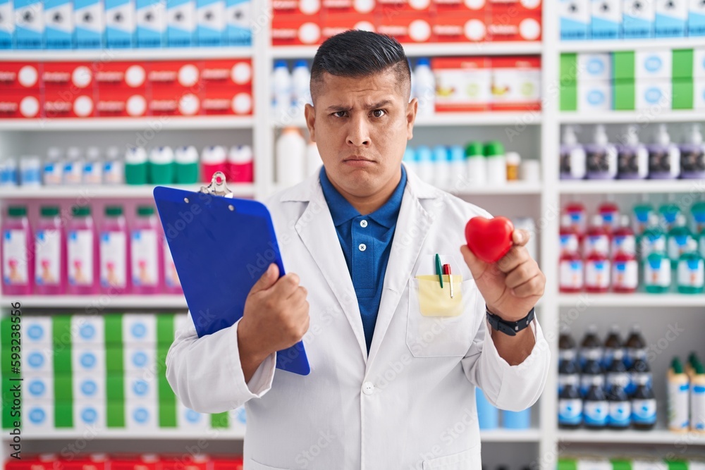 Young hispanic man working at pharmacy drugstore holding heart skeptic and nervous, frowning upset because of problem. negative person. Young hispanic man working at pharmacy drugstore holding heart skeptic and nervous, frowning upset because of problem. negative person.