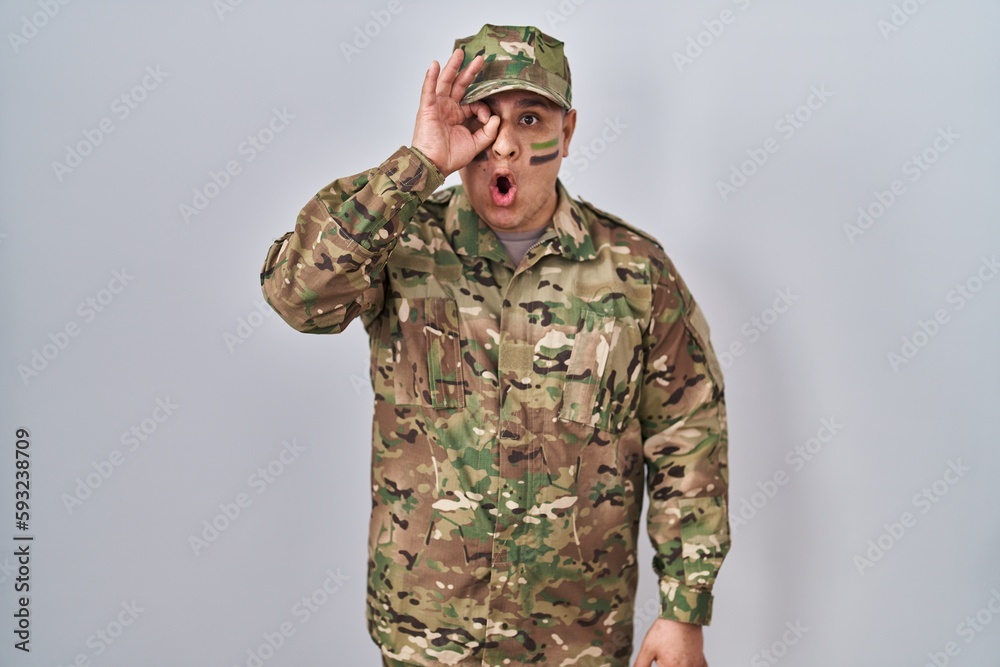 Hispanic young man wearing camouflage army uniform doing ok gesture shocked with surprised face, eye looking through fingers. unbelieving expression. Hispanic young man wearing camouflage army uniform doing ok gesture shocked with surprised face, eye looking through fingers. unbelieving expression.