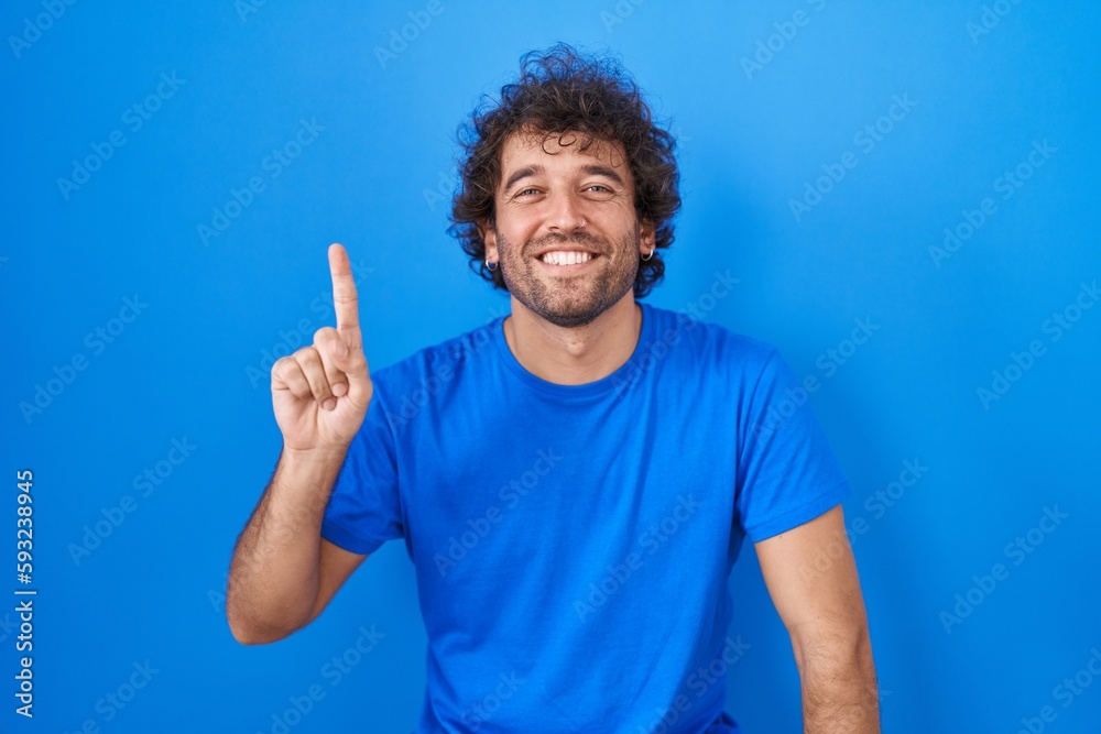 Naklejka premium Hispanic young man standing over blue background showing and pointing up with finger number one while smiling confident and happy.