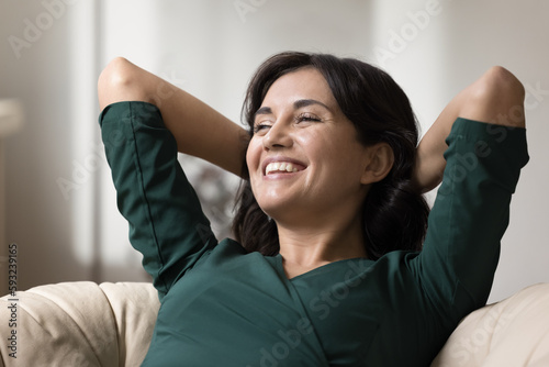 Close up happy young 30s woman put hands behind head resting seated in comfortable chair, enjoy relaxation inside modern sunny living room with air conditioning system. Leisure, pastime, stress-free