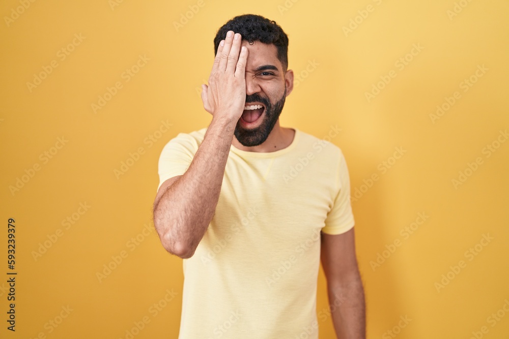 Hispanic man with beard standing over yellow background covering one eye with hand, confident smile on face and surprise emotion. Hispanic man with beard standing over yellow background covering one eye with hand, confident smile on face and surprise emotion.