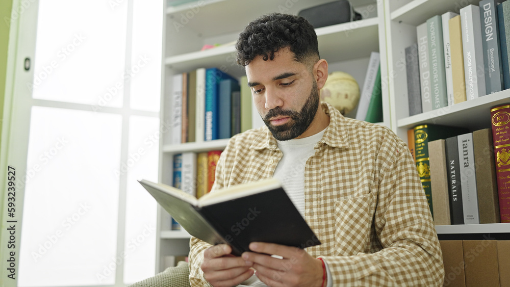 Fototapeta premium Young hispanic man student reading book at library university