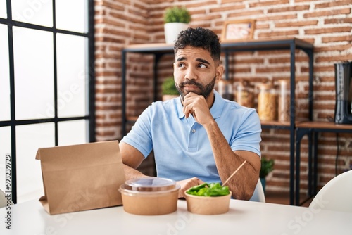 Hispanic man with beard eating delivery salad serious face thinking about question with hand on chin, thoughtful about confusing idea