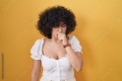 Young brunette woman with curly hair standing over yellow background feeling unwell and coughing as symptom for cold or bronchitis. health care concept.