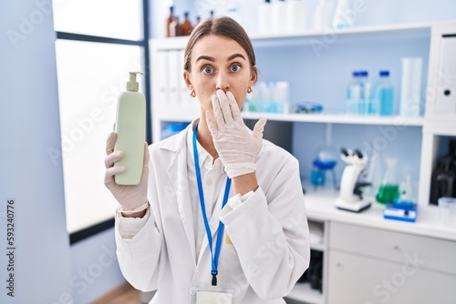 Young caucasian woman working at scientist laboratory holding body lotion covering mouth with hand  shocked and afraid for mistake. surprised expression