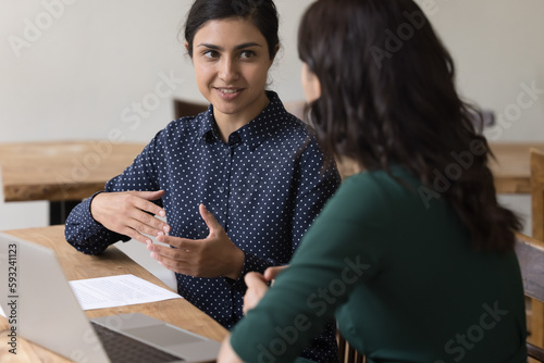 Two young businesswomen, colleagues or students discuss joint task seated at desk with laptop. Manager makes commercial offer, proposal, convinces customer to buy company services. Business meeting