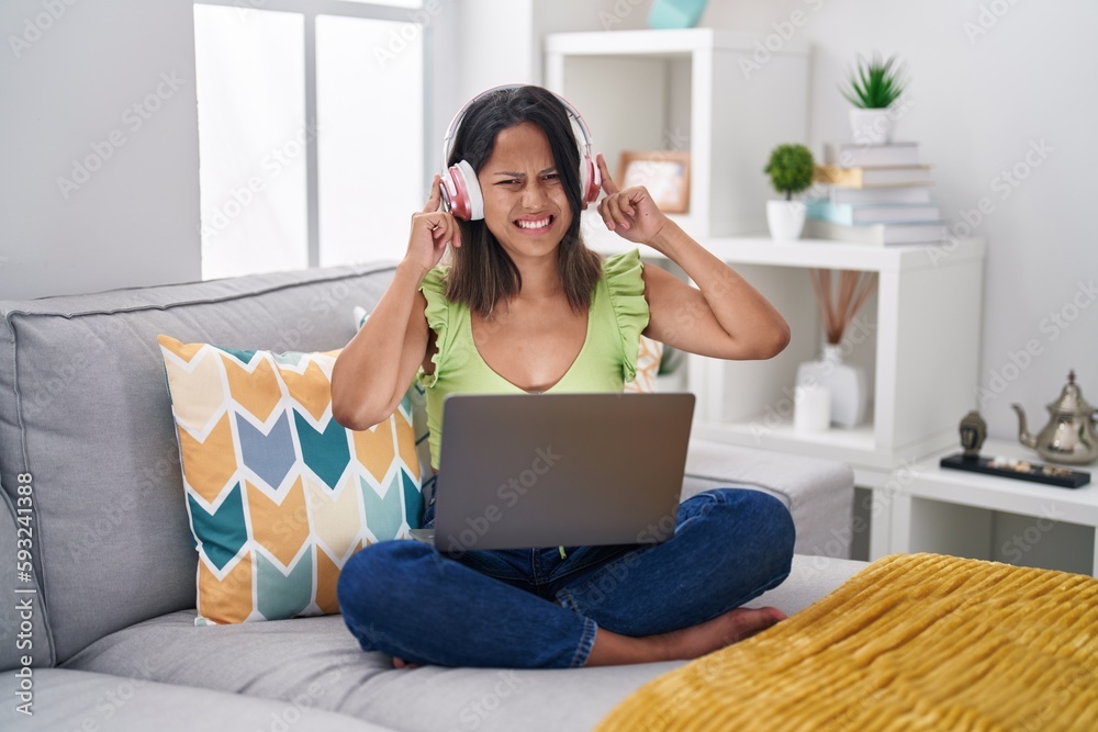 Hispanic young woman using laptop at home covering ears with fingers with annoyed expression for the noise of loud music. deaf concept. Hispanic young woman using laptop at home covering ears with fingers with annoyed expression for the noise of loud music. deaf concept.