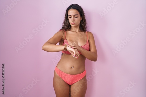 Young hispanic woman wearing lingerie over pink background checking the time on wrist watch, relaxed and confident