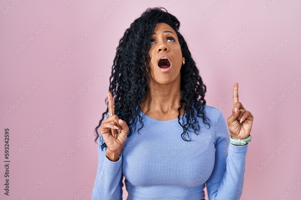 Middle age hispanic woman standing over pink background amazed and surprised looking up and pointing with fingers and raised arms. Middle age hispanic woman standing over pink background amazed and surprised looking up and pointing with fingers and raised arms.